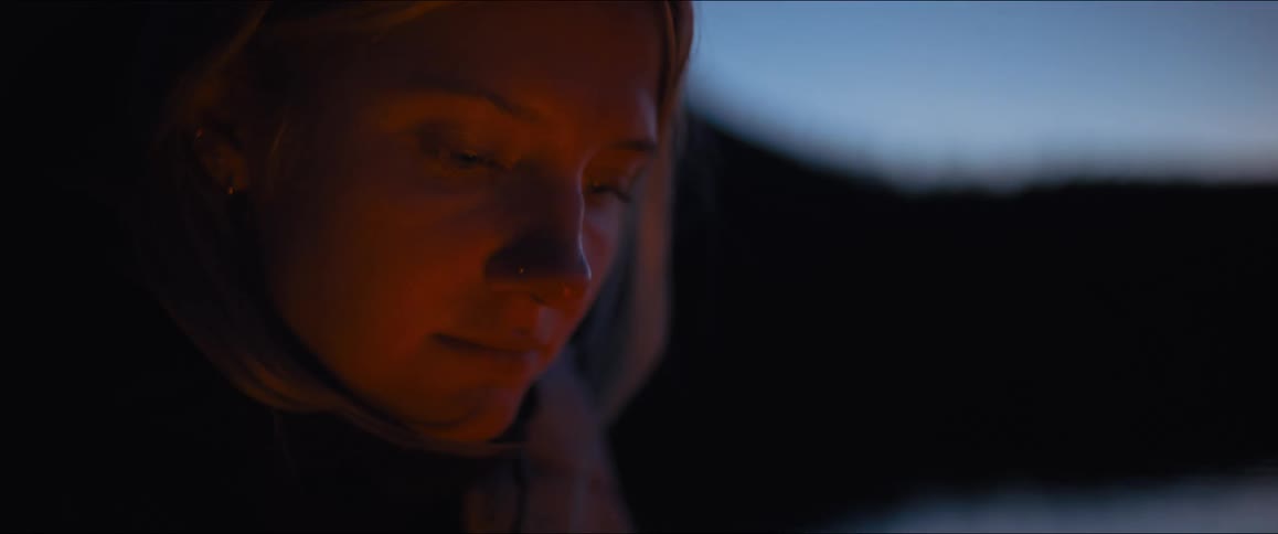 A tight shot shows a womans face in profile as she looks down at something out of frame the light of a fire also out of frame casts a warm glow on the side of her face