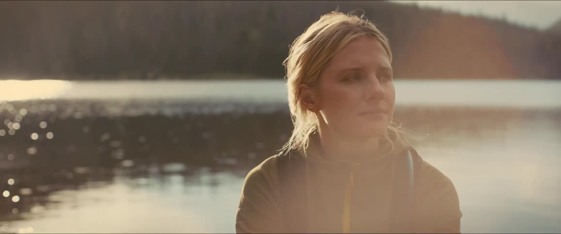 A female hiker seen in a halfbody shot is standing by a lake she has a pensive expression her gaze shifting from looking to the side to looking directly forward