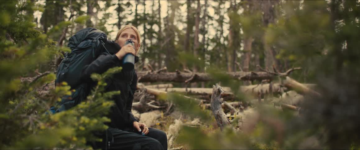 A female hiker wearing a large backpack sits down on a fallen log to take a drink from her water bottle she looks tired but content as she takes a break in the forest