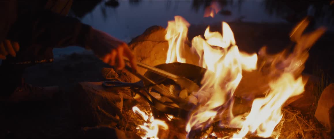 A person cooks food over a large campfire in the outdoors at night
