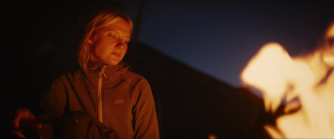 A womans face is illuminated by the warm glow of a campfire at night as she uses tongs to tend to a barbecue cooking a meal while camping outdoors