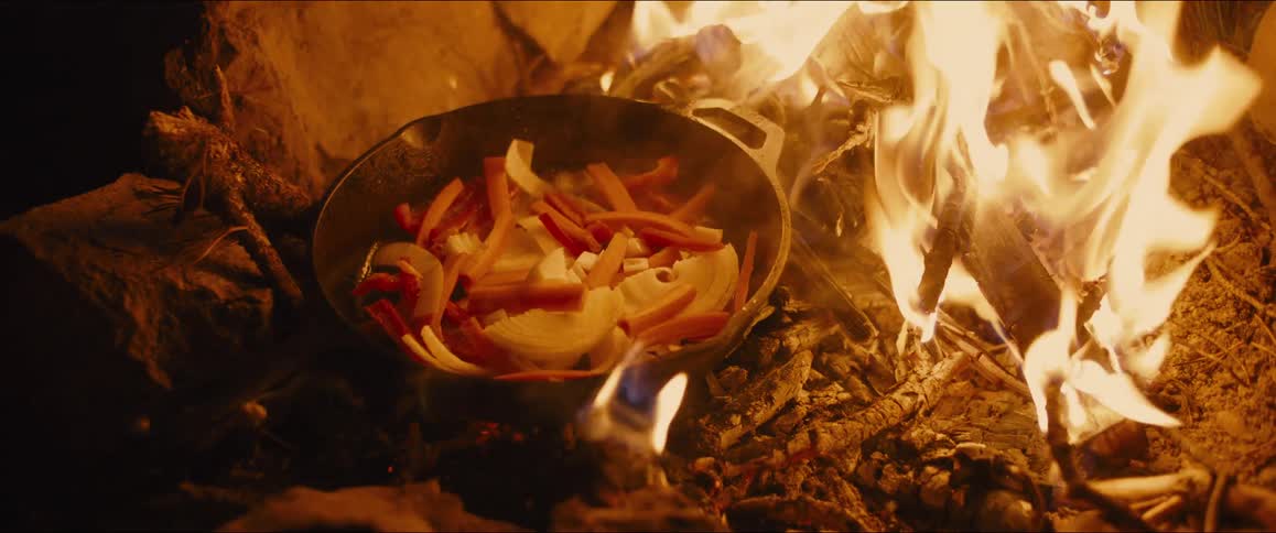 A small castiron pot is placed directly in the flames of a campfire and a pair of hands drops in some chopped vegetables the pot containing what looks like sliced onions and red peppers begins to sizzle and steam as it cooks over the open flame