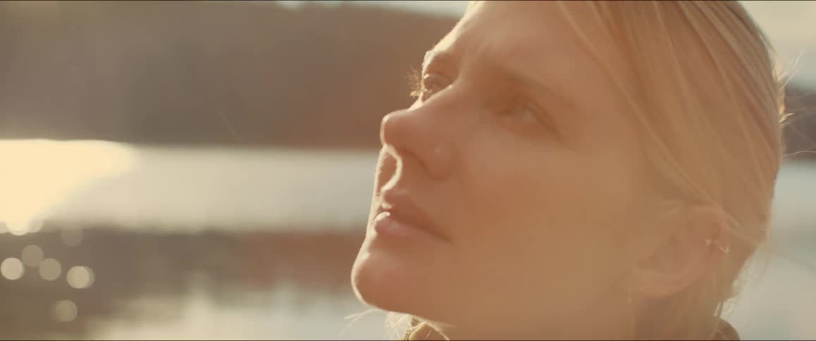 A blonde woman with her hair pulled back stands near a body of water illuminated by the golden light of the sun she slowly raises her head closing her eyes as she tilts her face up towards the sky appearing to enjoy the warmth and light
