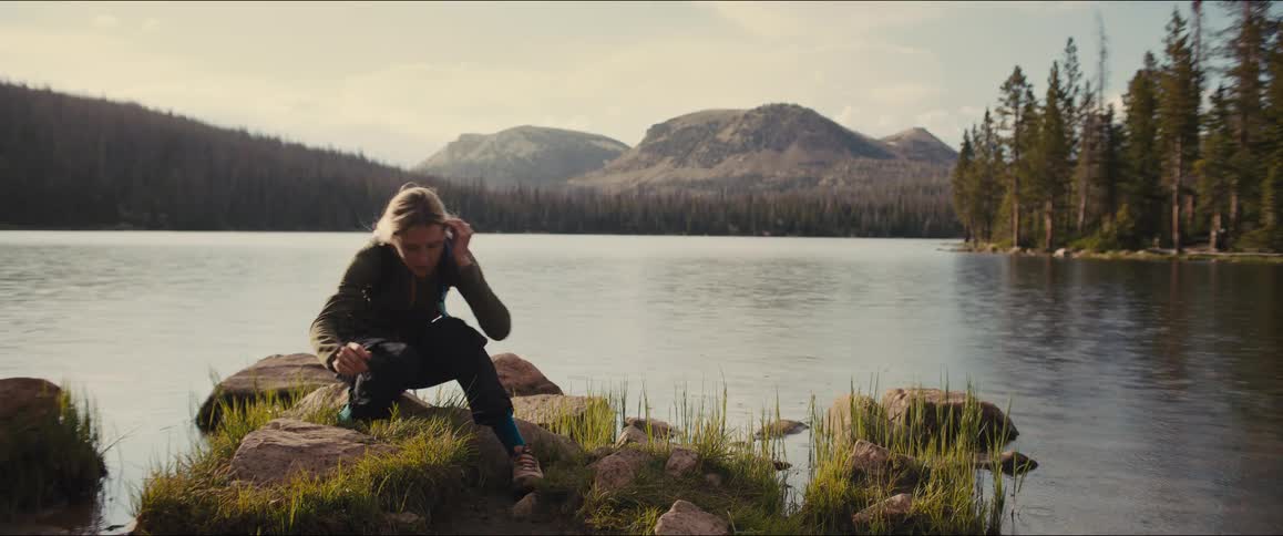 A person is sitting on rocks by a lake tying their shoe and then stands up to walk away from the camera