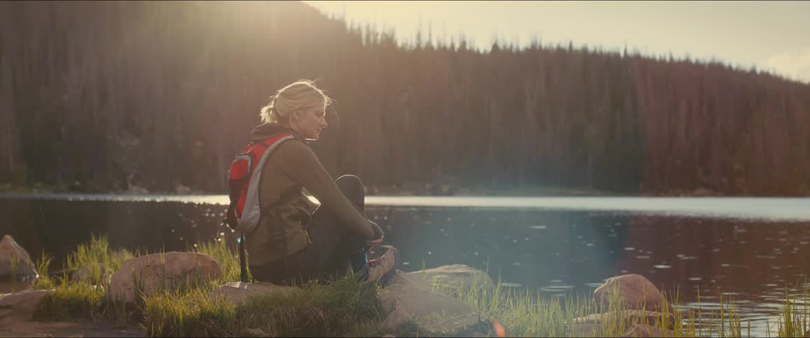 Woman sits on the shore of a lake while the sun shines over the forested mountain across the water