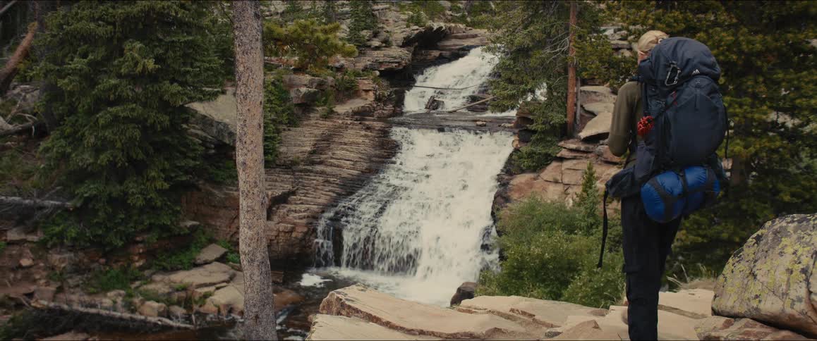 Female hiker walks to stand at a point overlooking a waterfall