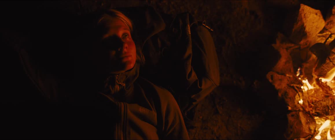 The woman is laying on the ground near the campfire at night