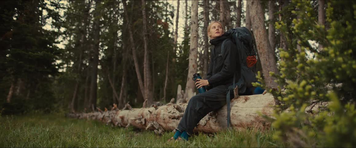 A blonde female hiker wearing a black jacket and a large backpack sits on a fallen tree trunk in a forest she drinks from a blue water bottle before looking up thoughtfully and then getting up to continue on her way