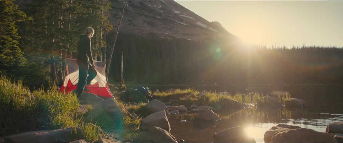 A person is setting up a tent next to a body of water with a mountain and a forest in the background