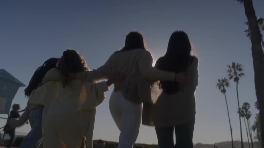 Four female friends walking side by side on a sidewalk at sunset hugging each other and having fun