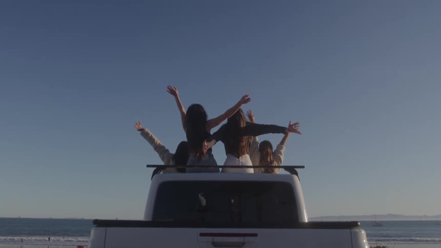 Four friends cheer with arms raised while sitting on a truck by the beach