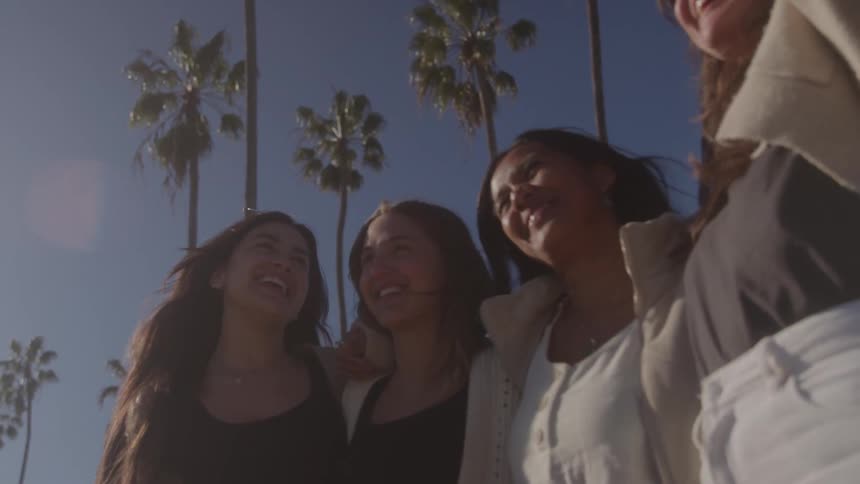 A lowangle shot captures a group of four diverse women standing together smiling and laughing palm trees are silhouetted against a clear blue sky behind them