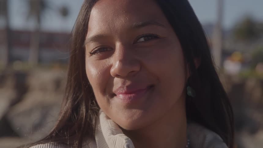 A closeup portrait shows a woman with long dark hair looking directly into the camera with a warm gentle smile on a sunny day outdoors