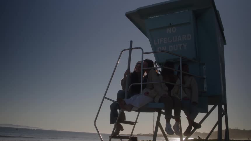 Female friends sit on the platform of a lifeguard stand during a dimly lit day at the beach