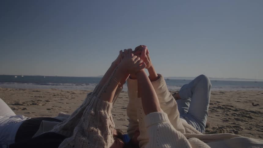 Three people are lying on the sand and raising their hands to meet in the middle while facing the ocean