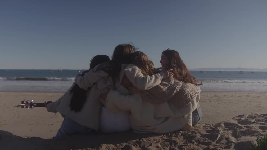 A group of four young women with long hair are sitting together on a sandy beach they have their arms around each others shoulders facing the ocean with a guitar resting beside one of them