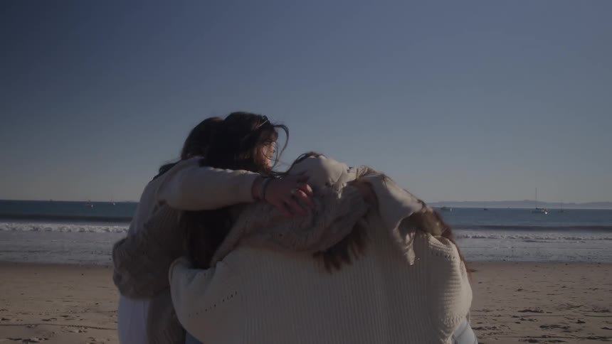On a sunny beach two women hugging are joined by two more friends and the group of four walks and skips joyfully towards the ocean together in a big hug