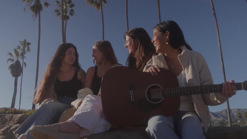 Four smiling friends sitting outdoors with one of them holding a guitar