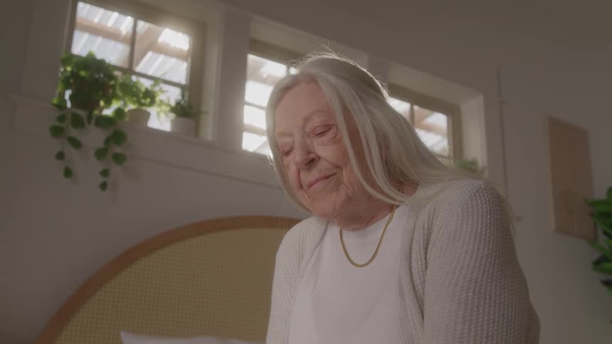 An elderly woman with long white hair sits in a brightly lit room with a peaceful expression she is wearing a white sweater and a gold necklace