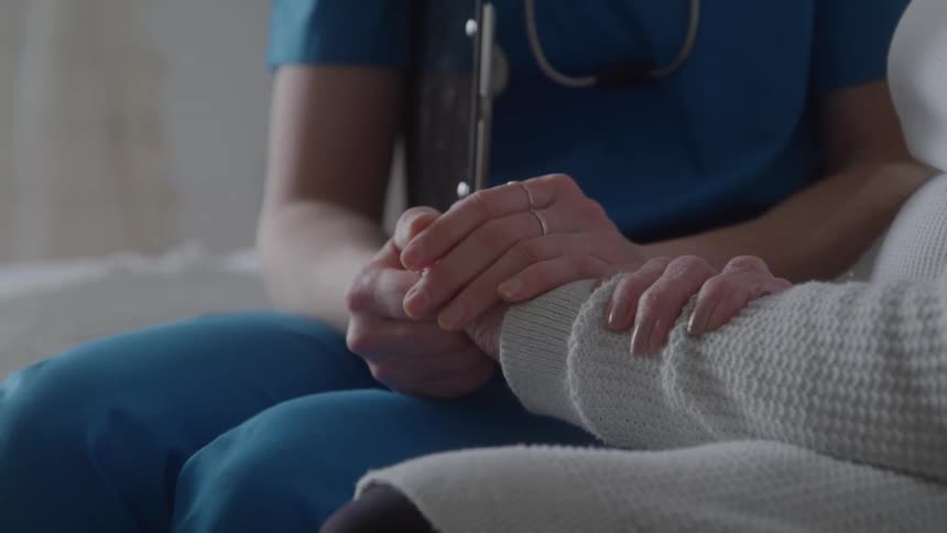 A medical professional dressed in blue scrubs is holding the hand of an elderly patient who is wearing a soft gray sweater this gesture conveys comfort and emotional support during a medical consultation or visit