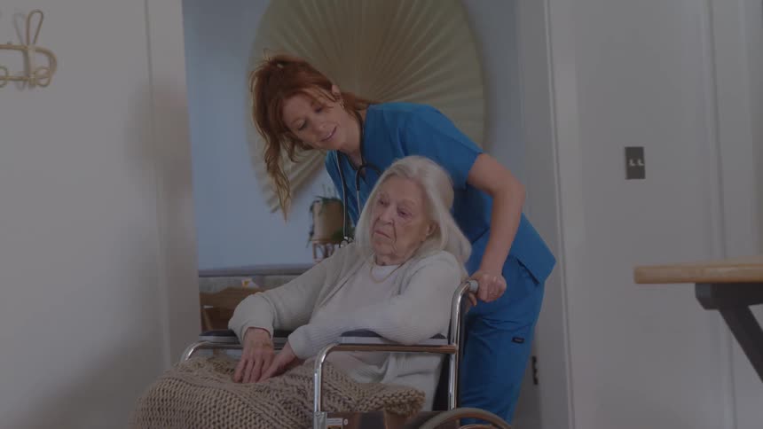 A compassionate female nurse in blue scrubs smiles and talks with an elderly woman while pushing her in a wheelchair through her home
