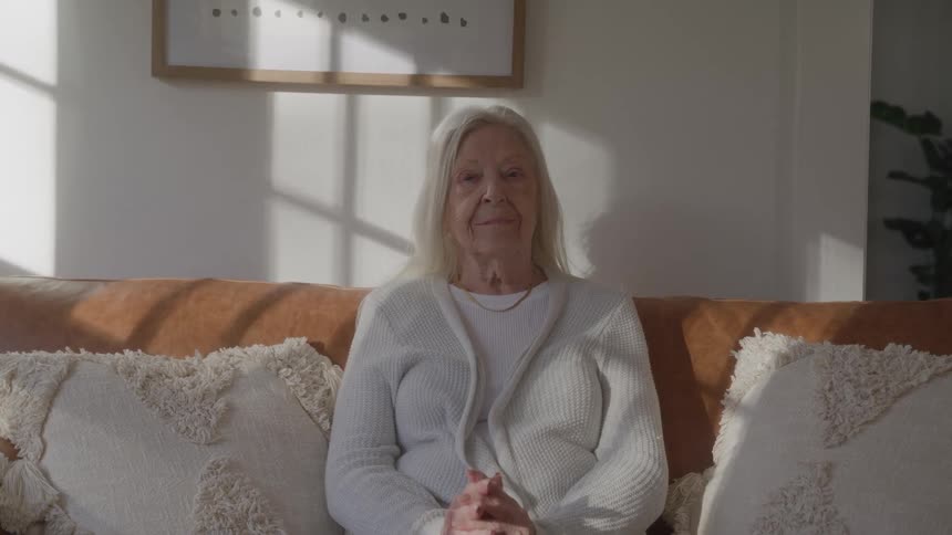 The camera slowly moves closer to an elderly woman with long white hair who sits calmly with her hands clasped on a leather sofa in a sunlit room while looking directly forward with a neutral expression