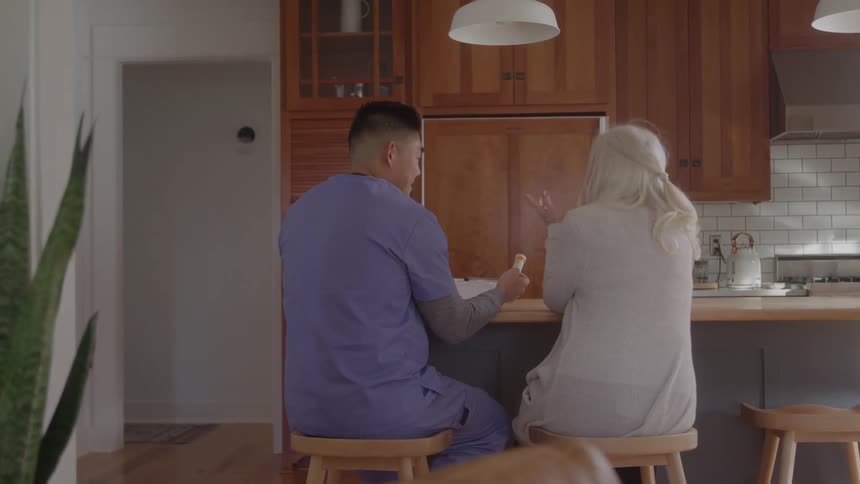 A male nurse in blue scrubs sits with an elderly woman at her kitchen island patiently explaining her medication or care plan to her
