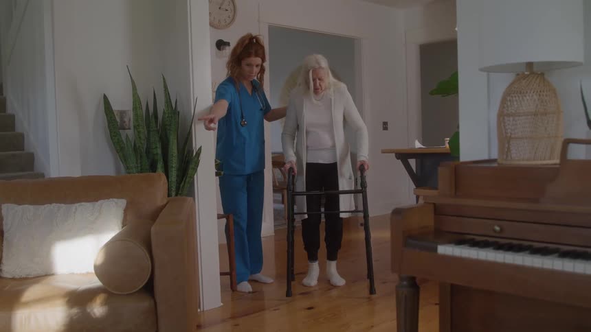 Nurse helping an elderly woman walk with a walker around the room in her house