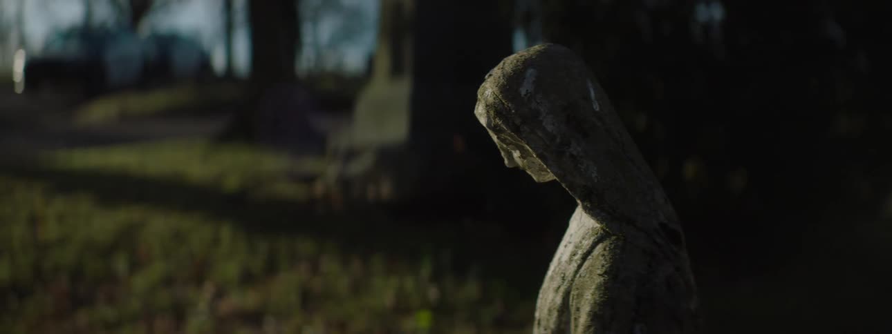 A shot of a statue in a cemetery pans out to show a landscape with a bright sun in the background