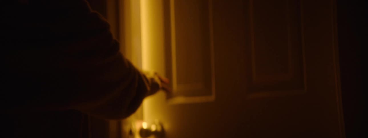 A dark shot of a closed door with a bright streak of light peeking through the crack a hand hesitantly reaches out placing a palm on the doorframe as if preparing to step through