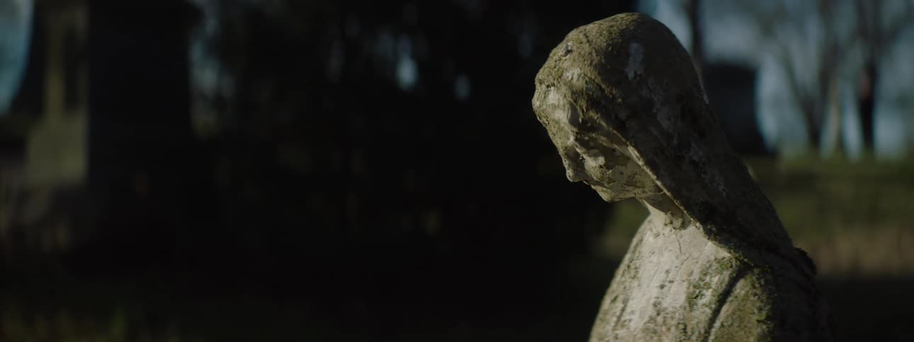 Statue of a female figure standing in front of a tombstone in a cemetery at midday
