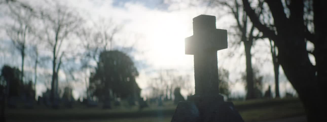 A cemetery with a stone cross gravestone in the foreground silhouetted against a cloudy sky and bare winter trees the lighting creates a somber reflective atmosphere