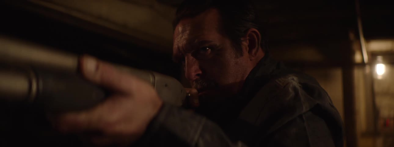 A shot shows an armed man in a dimly lit room pointing a shotgun he peeks cautiously from behind a wall his face tense and focused on an offscreen threat