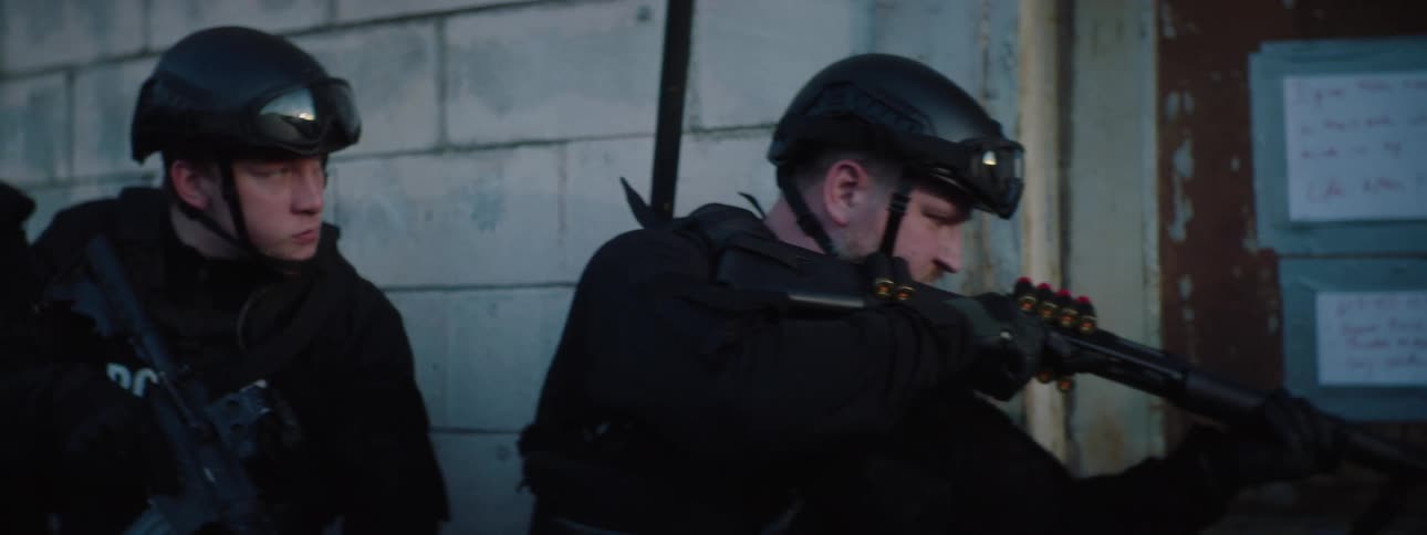 Two male police officers in black tactical gear and helmets crouch next to a stone wall the one on the left who is african american holds a rifle while the one on the right who is caucasian is seen holding and loading a shotgun
