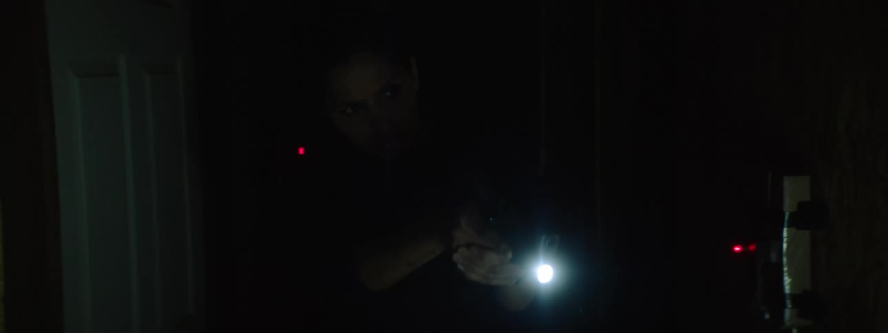 A video game scene of a focused woman in tactical gear aiming her flashlight through the darkness