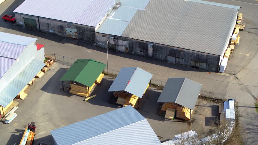 An aerial view shows a lumber yard with multiple warehouses and covered storage areas in daylight