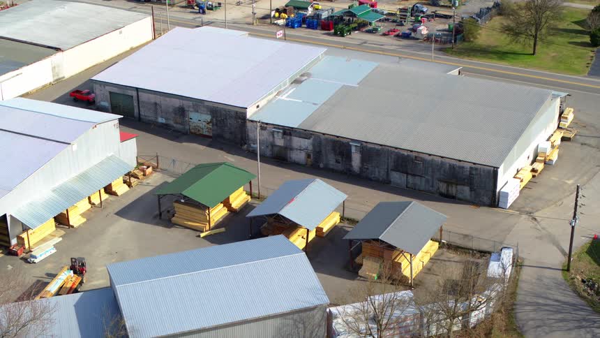 A slow orbiting aerial shot circles over a lumber yard revealing large warehouses neatly stacked lumber and several prefabricated sheds on display
