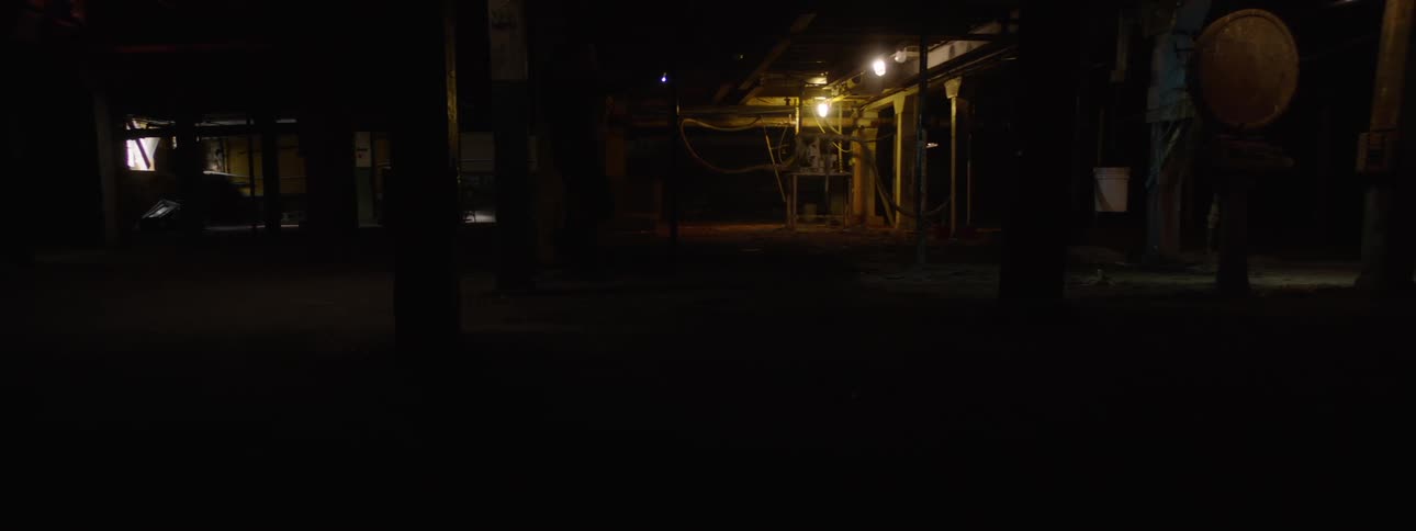 In the video the interior of a dimly lit dark warehouse or industrial space is shown a person walks through the background and as they pass a bright light turns on briefly illuminating the area