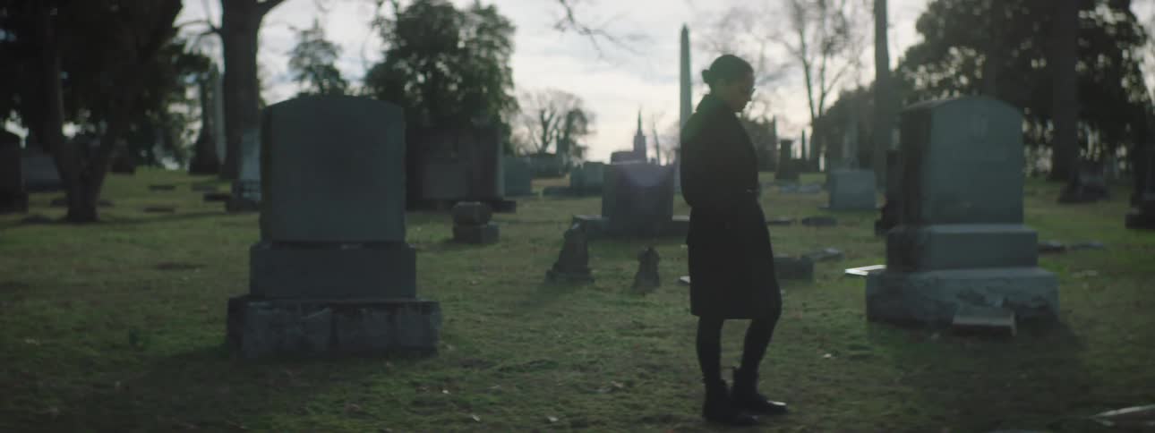 Young woman with a ponytail wearing black clothes standing in a graveyard at midday with a sad expression
