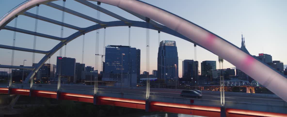 An aerial shot follows a car driving over a bridge at dusk revealing the illuminated nashville skyline in the background