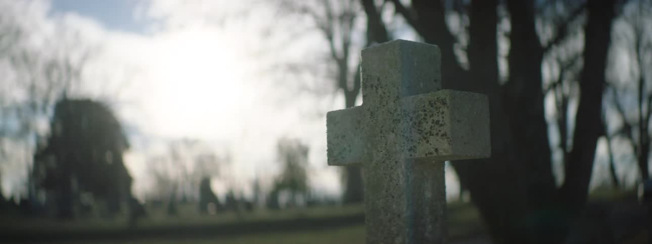 A weathered gray stone cross in a cemetery is shown in a closeup shot the camera slowly pans across the cross and the surrounding bare trees with the sunlight creating a bright lens flare