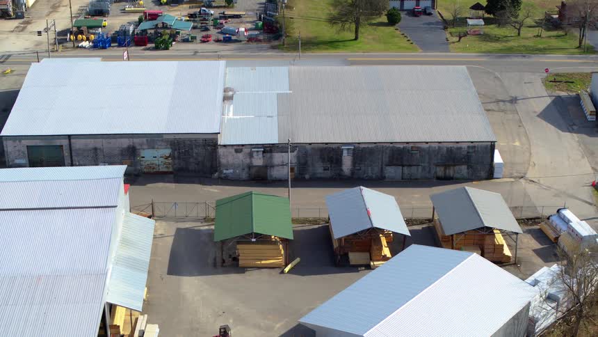 A lumber yard with large warehouses and smaller sheds filled with stacked wood adjacent to a road with distant traffic