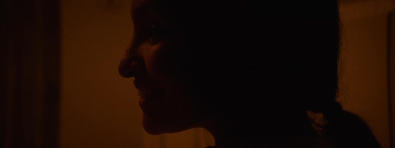 In the video the profile of a womans head is shown with the right side of her face illuminated by a soft orange light her expression changes as she smiles and appears to be speaking to someone offscreen