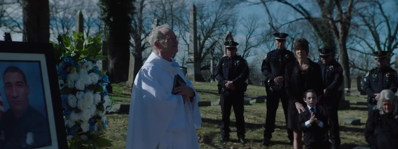 A priest speaks at a somber graveside funeral for a fallen police officer as the camera pans past grieving family and uniformed officers to rest on the casket