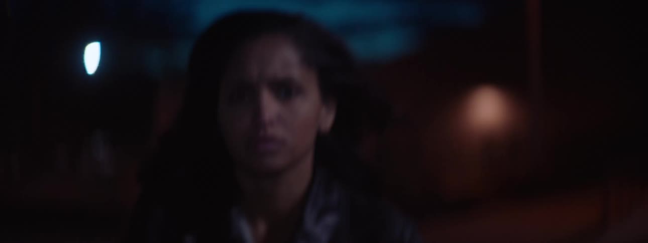 In a shaky handheld shot a woman with a worried expression walks at a fast pace down a dark city street at night her face lit by ambient streetlights