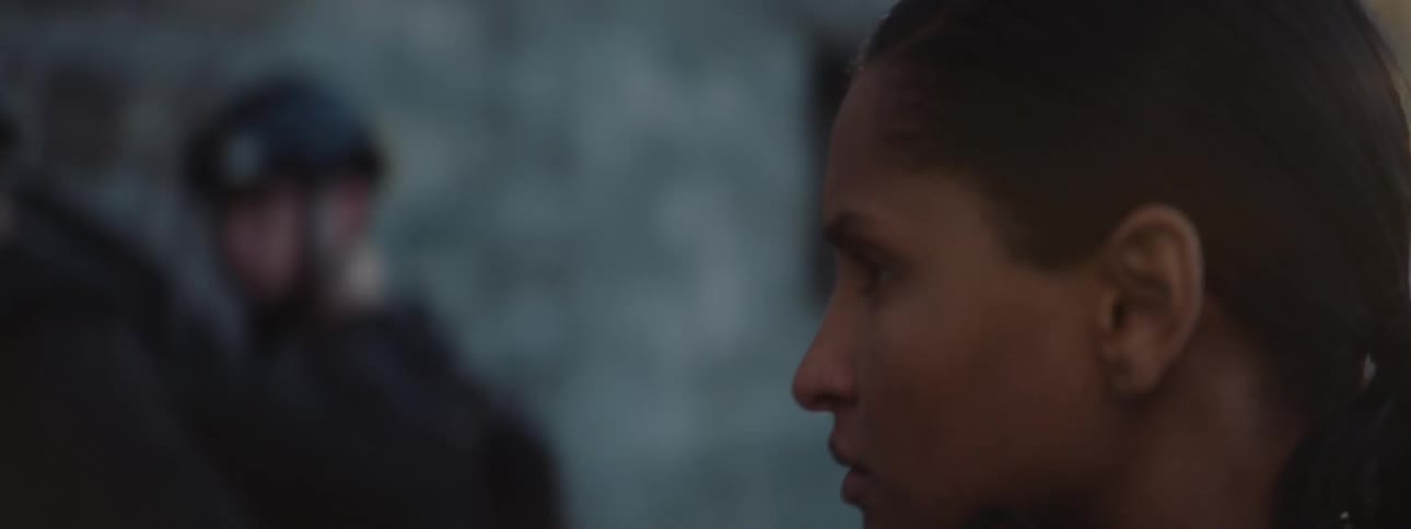In the video a woman with a ponytail and a serious expression is seen standing outdoors in a tactical vest she is holding and manipulating a black pistol