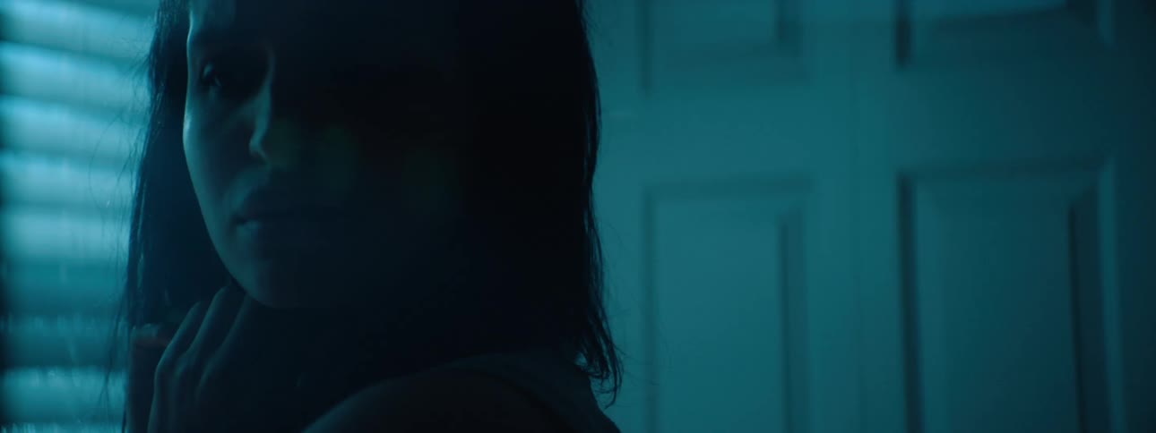 A woman looks tense and wary in dim blue light half her face obscured by shadow