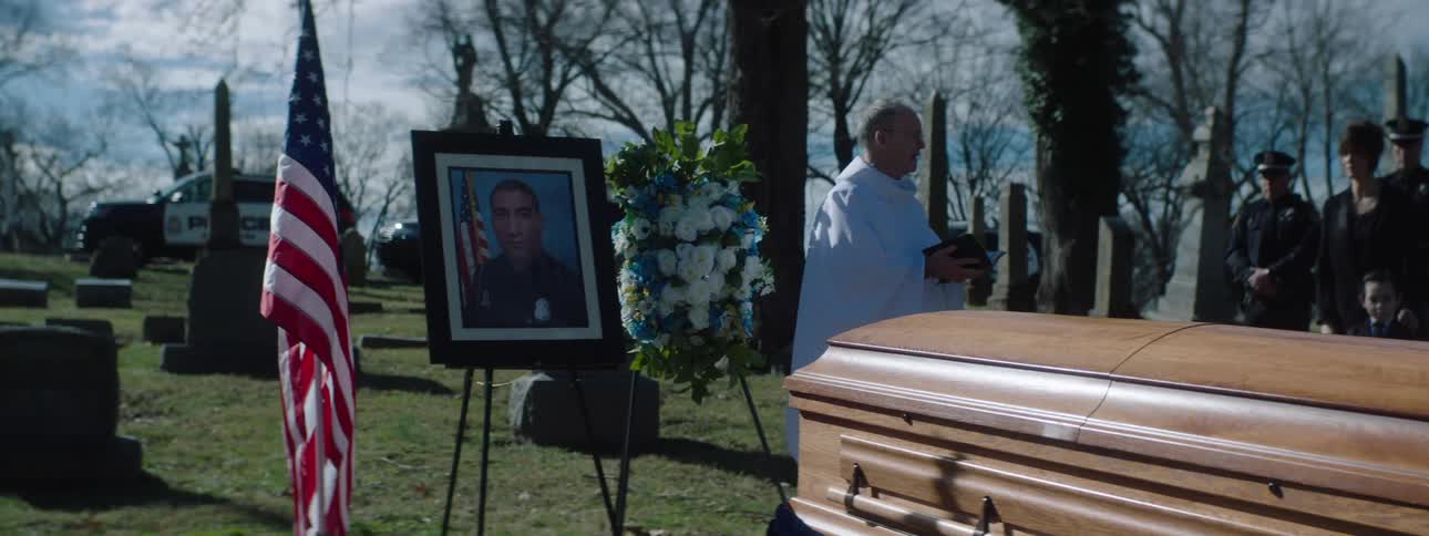 At a somber graveside funeral for a police officer a priest officiates beside the casket and a framed portrait while fellow officers and family look on
