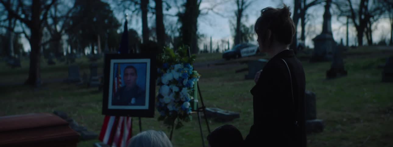 People are gathered in a cemetery around a coffin and a framed photograph on an easel and a woman is looking at the photograph