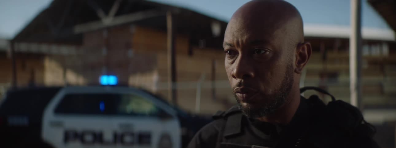 A police officer with a worried expression is standing in an outdoor setting with a police car in the background